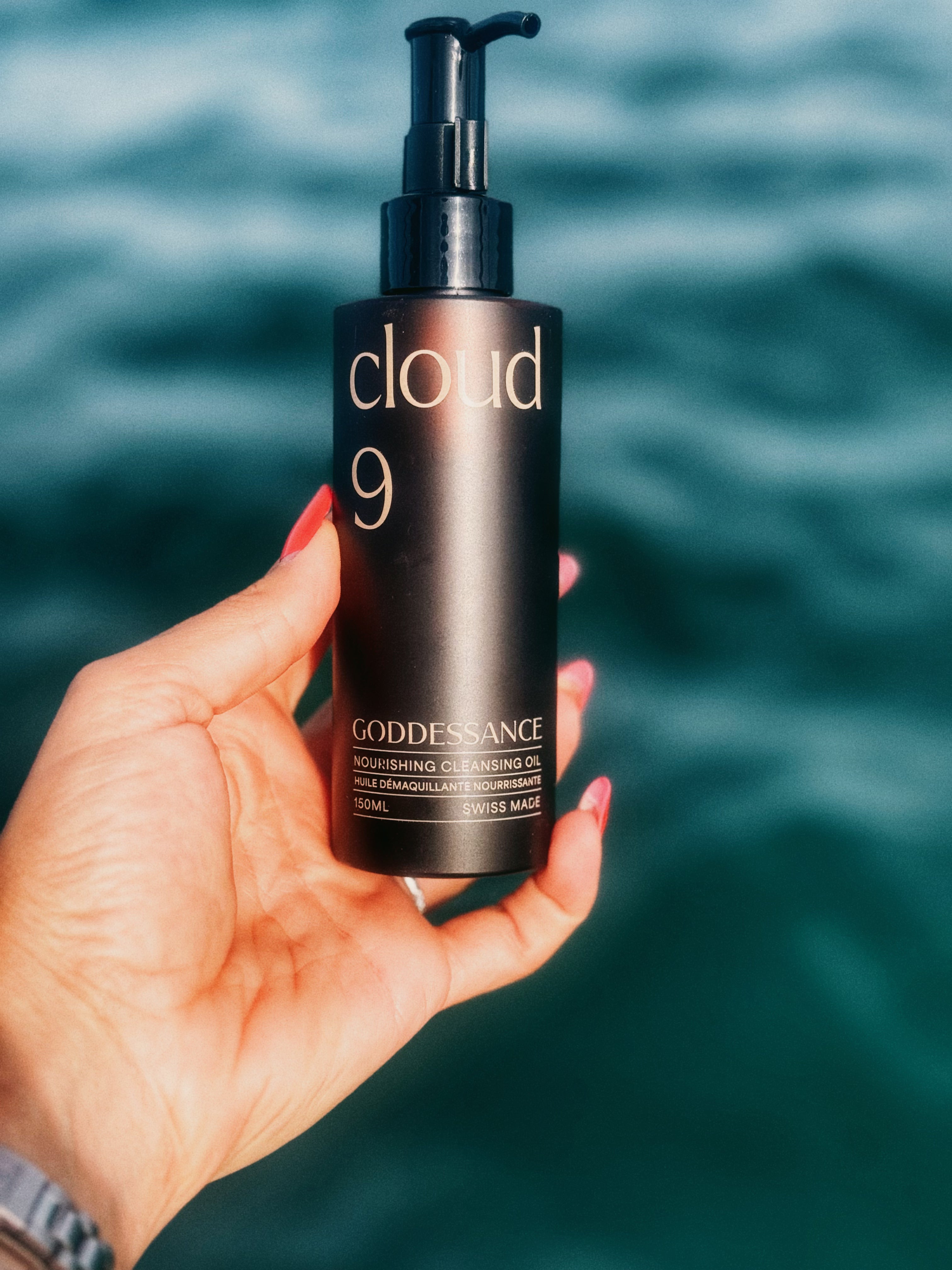 Hand holding a Cloud 9 cleansing oil bottle against a blue sea backdrop