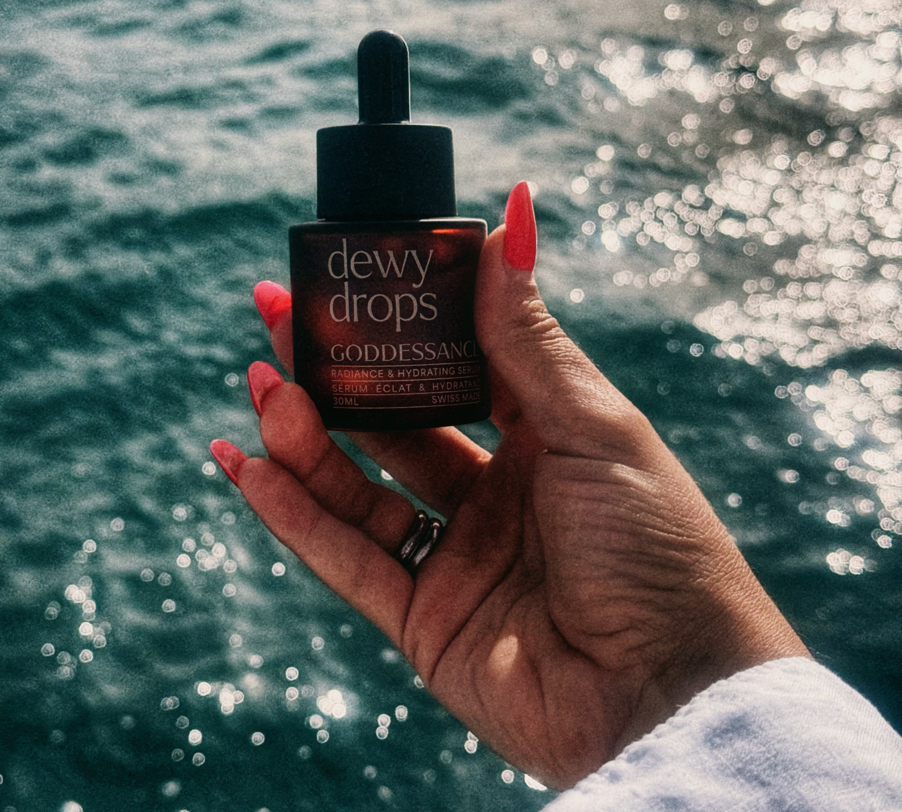 Hand holding a Dewy Drops serum bottle against a sparkling sea